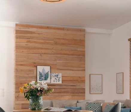 Eine runde, geflochtene Deckenlampe mit sichtbaren Glühbirnen beleuchtet ein modernes Wohnzimmer. Im Vordergrund steht ein runder Holztisch mit einer Vase bunter Blumen und einer offenen Zeitschrift, flankiert von zwei grauen Stühlen. Eine Holzakzentwand und ein graues Ecksofa mit Zierkissen sind im Hintergrund.