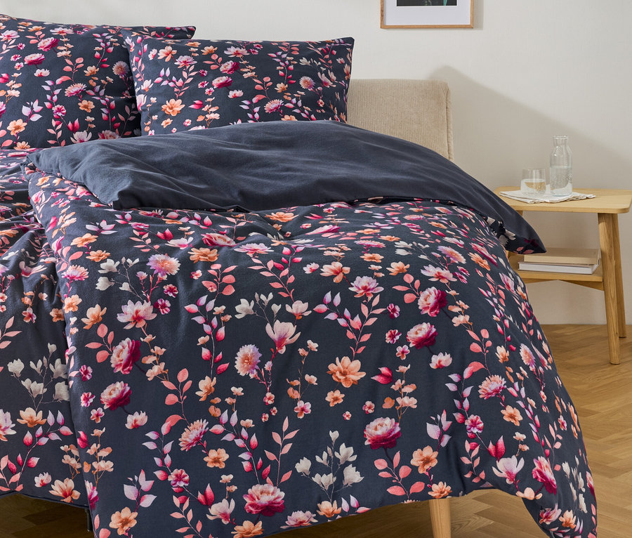 Bett mit dunkelblauer Bettwäsche mit Blumenmuster in Pink, Orange und Weiß. Auf dem Nachttisch stehen eine Wasserflasche und ein Glas. An der Wand hängen ein Bild und eine rosa Pendelleuchte.