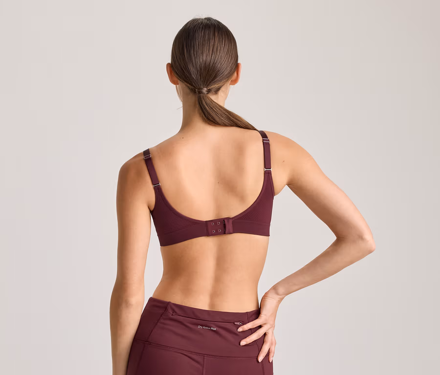 Frau von hinten, trägt einen bordeauxroten Sport-BH und passende Leggings. Ihr Haar ist zu einem Pferdeschwanz gebunden, ihre linke Hand liegt auf ihrer Hüfte.