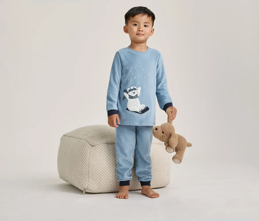 Kleiner Junge in blauem Pyjama mit Eisbär-Motiv, der einen braunen Stoffhund hält und neben einem hellen Pouf steht.