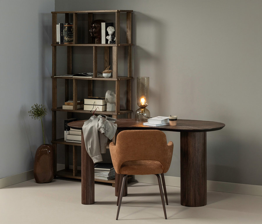 Ein dunkles Holzregal mit Büchern und Dekoartikeln steht in einer Zimmerecke. Daneben befindet sich ein ovaler Holzschreibtisch mit einem grauen Hemd, Büchern, einer Tasse und einer Glaslampe. Ein brauner Polsterstuhl steht vor dem Schreibtisch, und eine Vase mit einer Blume steht am Boden.
