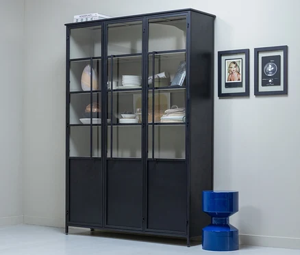 Ein schwarzer Metallschrank mit Glastüren, gefüllt mit weißem Geschirr und Textilien, steht vor einer hellgrauen Wand. Rechts davon hängen zwei gerahmte Bilder über einem leuchtend blauen, sanduhrförmigen Hocker.