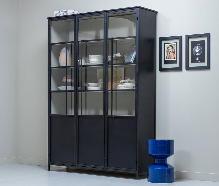 Ein schwarzer Metallschrank mit Glastüren, gefüllt mit weißem Geschirr und Textilien, steht vor einer hellgrauen Wand. Rechts davon hängen zwei gerahmte Bilder über einem leuchtend blauen, sanduhrförmigen Hocker.