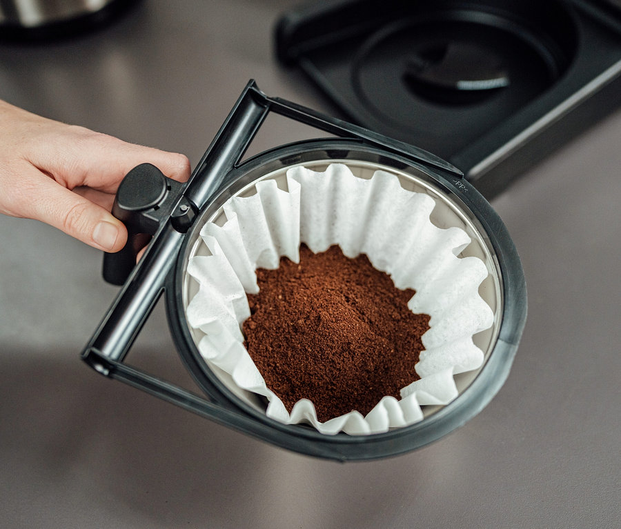 Hand hält Kaffeefilter mit Papierfilter und Kaffeepulver. Zu sehen ist eine Profi-Filterkaffeemaschine »Bravilor-Bonamat TH«, ohne Kanne.
