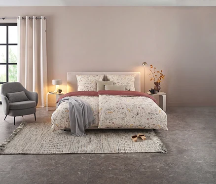 Gemütliches Schlafzimmer mit einem Bett, das mit floraler SCHÖNER WOHNEN Perkal-Bettwäsche bezogen ist, einem grauen Sessel und einem gestreiften Teppich.