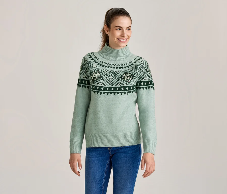 Lächelnde Frau in einem hellgrünen Rollkragenpullover mit dunkelgrünem und weißem Fair-Isle-Muster und blauen Jeans.