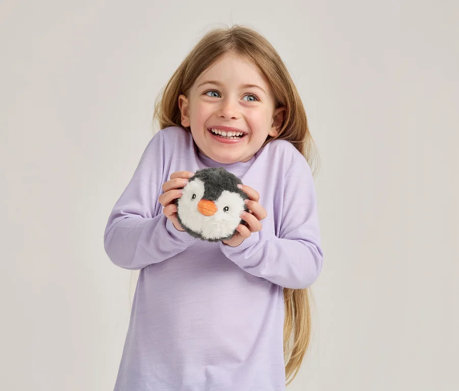 Ein lächelndes junges Mädchen mit langen blonden Haaren und blauen Augen, das ein hellviolettes Langarmshirt trägt und ein rundes Plüschpinguin-Spielzeug hält.