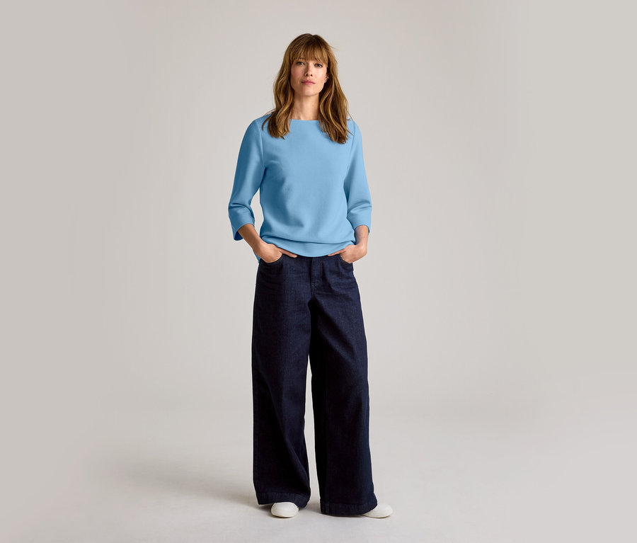 Frau mit schulterlangem Haar, die ein hellblaues Oberteil, eine dunkelblaue weite Jeans und weiße Leder-Sneaker trägt.