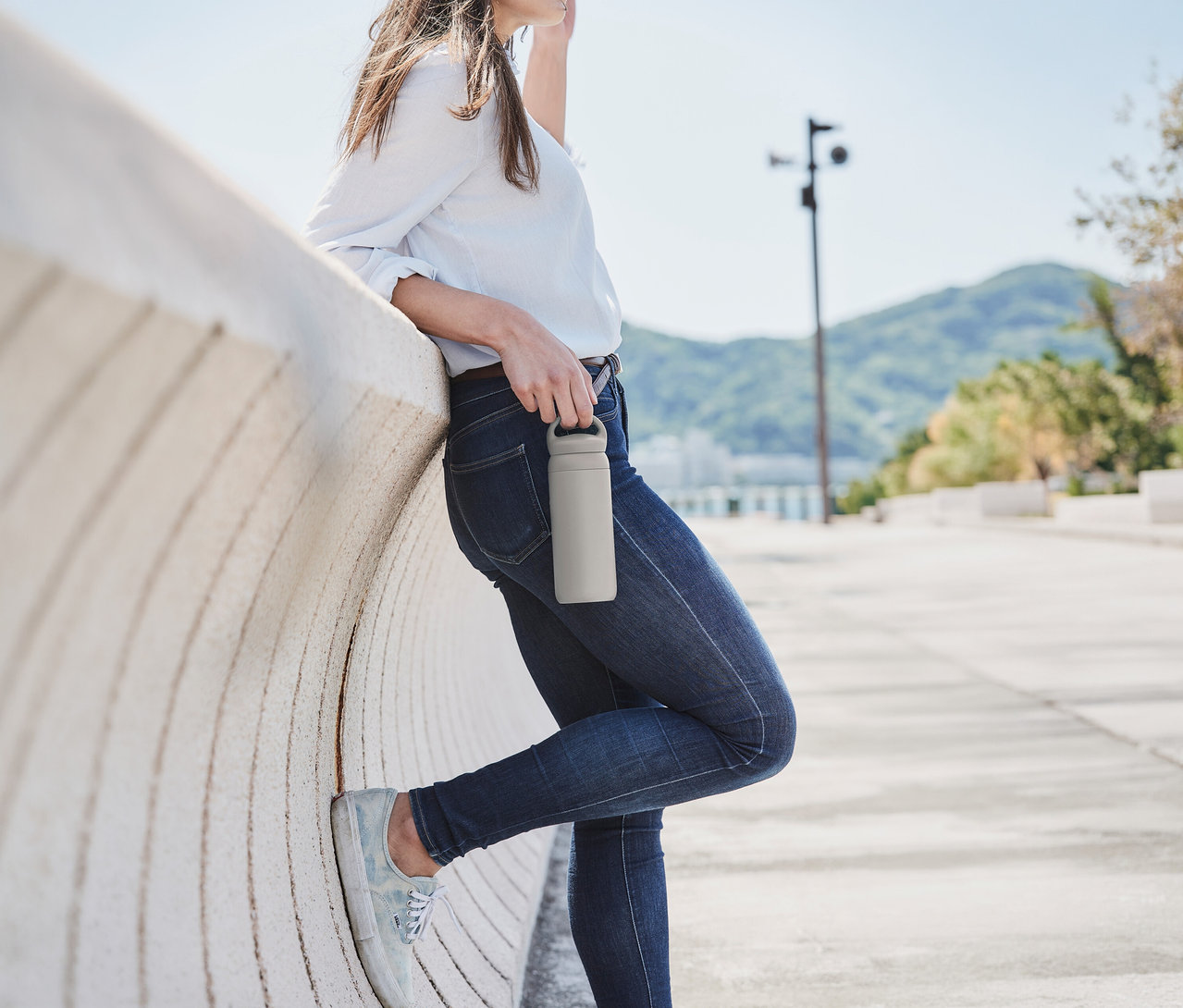 Eine Frau lehnt an einer Wand und hält einen beigen KINTO Tumbler »Day Off« in der Hand. Sie trägt Jeans und ein helles Hemd, im Hintergrund sind Hügel.