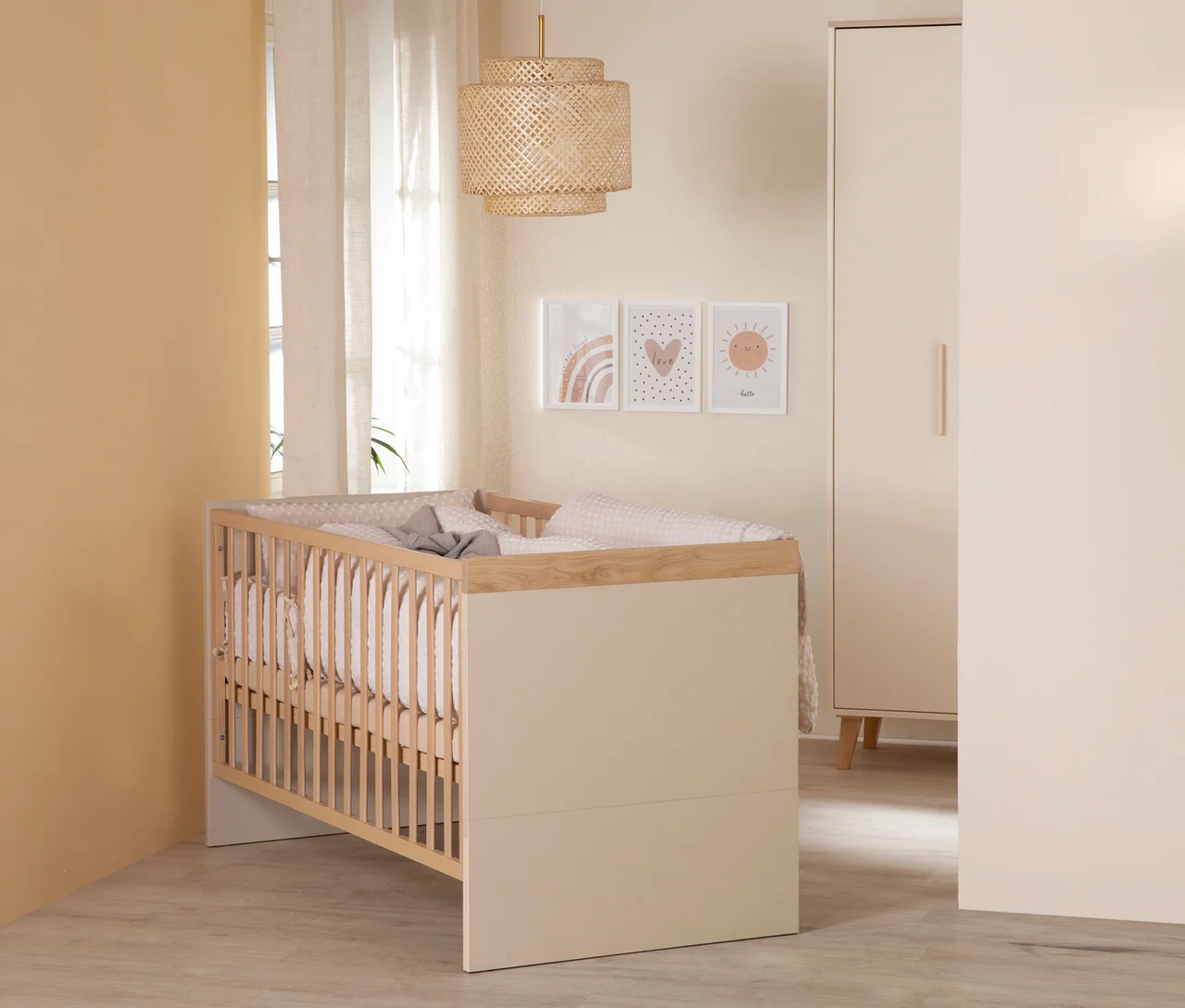 Ein neutrales Babyzimmer mit einem hellen Holz- und beigefarbenen Gitterbett, rosa gepunkteter Bettwäsche und grauer Decke. Rechts steht ein beigefarbener Kleiderschrank. Über dem Bett hängen drei Bilder mit Regenbogen, Herz und Sonne. Eine geflochtene Pendelleuchte und transparente Vorhänge am Fenster ergänzen den Raum.