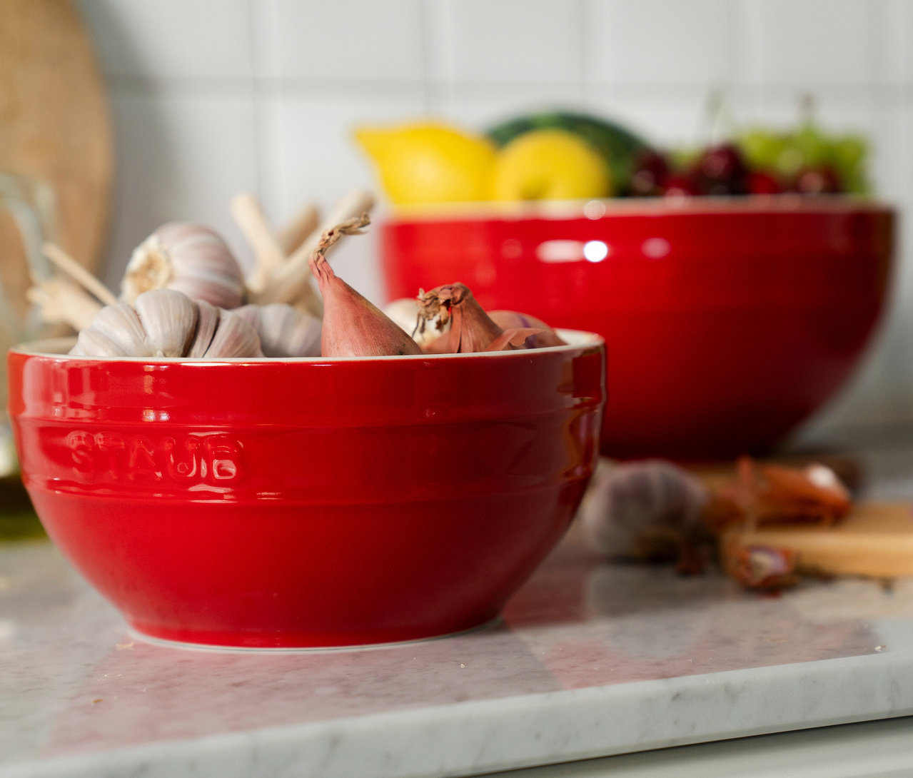 Kirschrote STAUB Keramik-Schüssel mit Knoblauch und Schalotten auf Marmor-Arbeitsplatte. Im Hintergrund weitere rote Schüssel mit Obst.