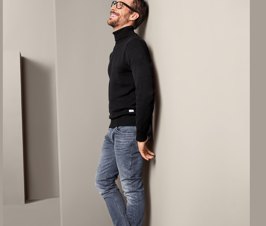 Mann lehnt an Wand, trägt Rollkragenpullover, Jeans und Turnschuhe.