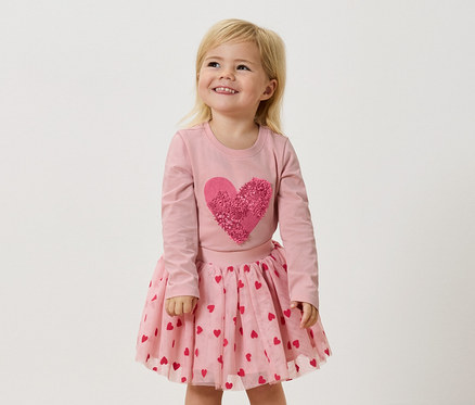 Ein kleines Mädchen mit blonden Haaren trägt ein rosa Langarmshirt mit einem großen, rosafarbenen Herz und einen rosa Tüllrock mit roten Herzen.