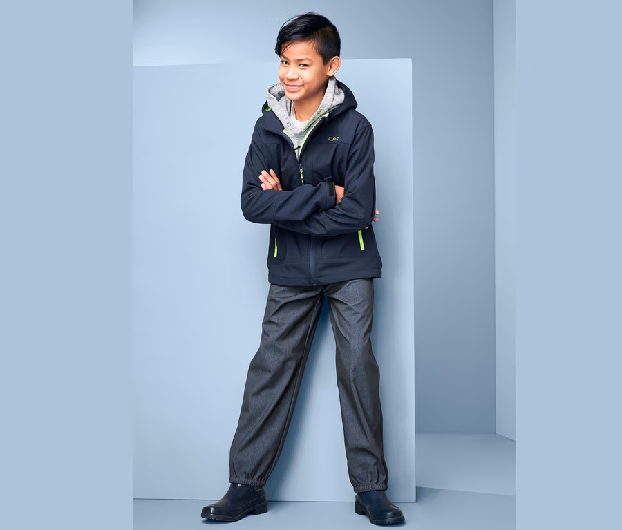 Ein Junge steht mit verschränkten Armen und trägt eine Softshellhose im Jeans-Look, eine Jacke und einen Hoodie.