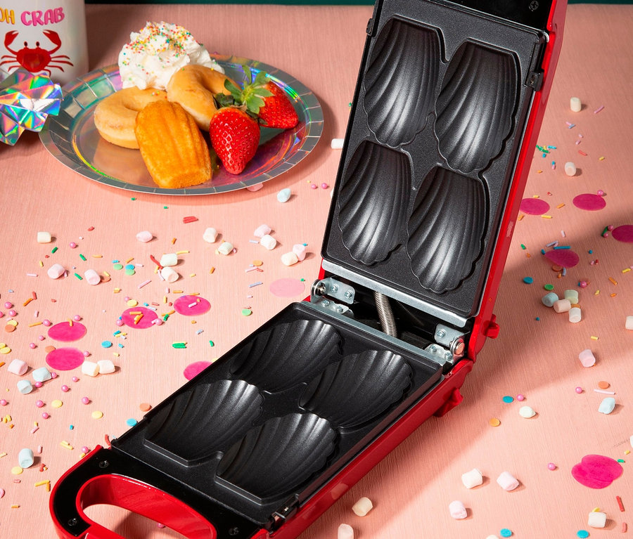 Ein offener, roter 6-in-1-Snackmaker auf rosafarbenem Hintergrund mit Konfetti, Teller mit Donuts, Erdbeeren und Madeleines.