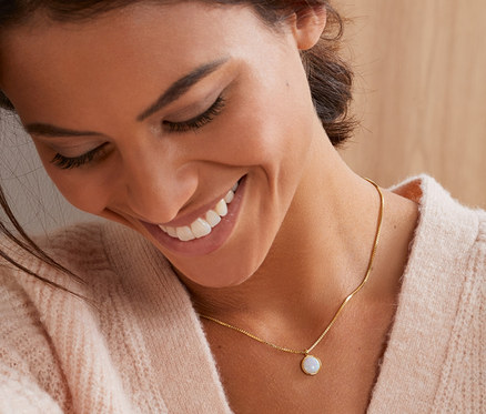 Lächelnde Frau berührt ihr Haar und trägt eine vergoldete Venezia-Kette mit einem Anhänger mit synthetischem Opal.