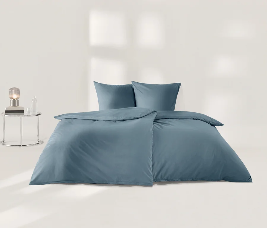 Blaue Perkal-Bettwäsche mit einem Bettbezug und zwei Kissenbezügen, ordentlich auf einem hellen Boden arrangiert, neben einem verchromten Beistelltisch mit Lampe und Glasflasche.