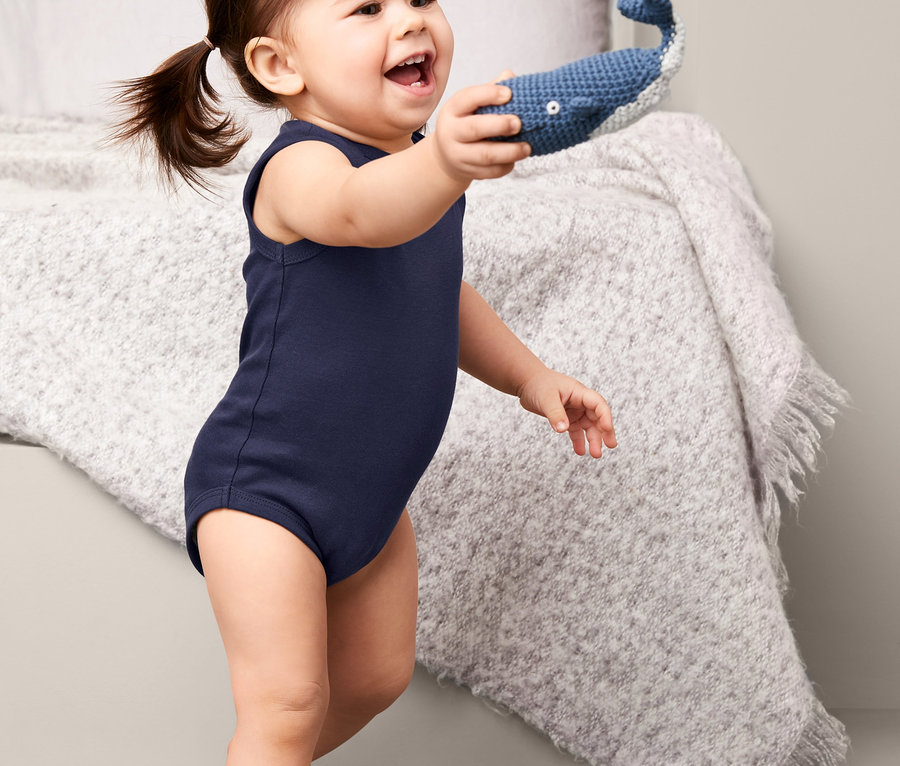 Ein Baby in einem marineblauen, ärmellosen Body hält ein blaues, gehäkeltes Walspielzeug.