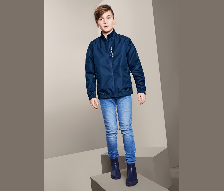 Ein Junge posiert in einer Allwetterjacke im Bomberjacken-Stil, blauen Jeans und Gummistiefeln.