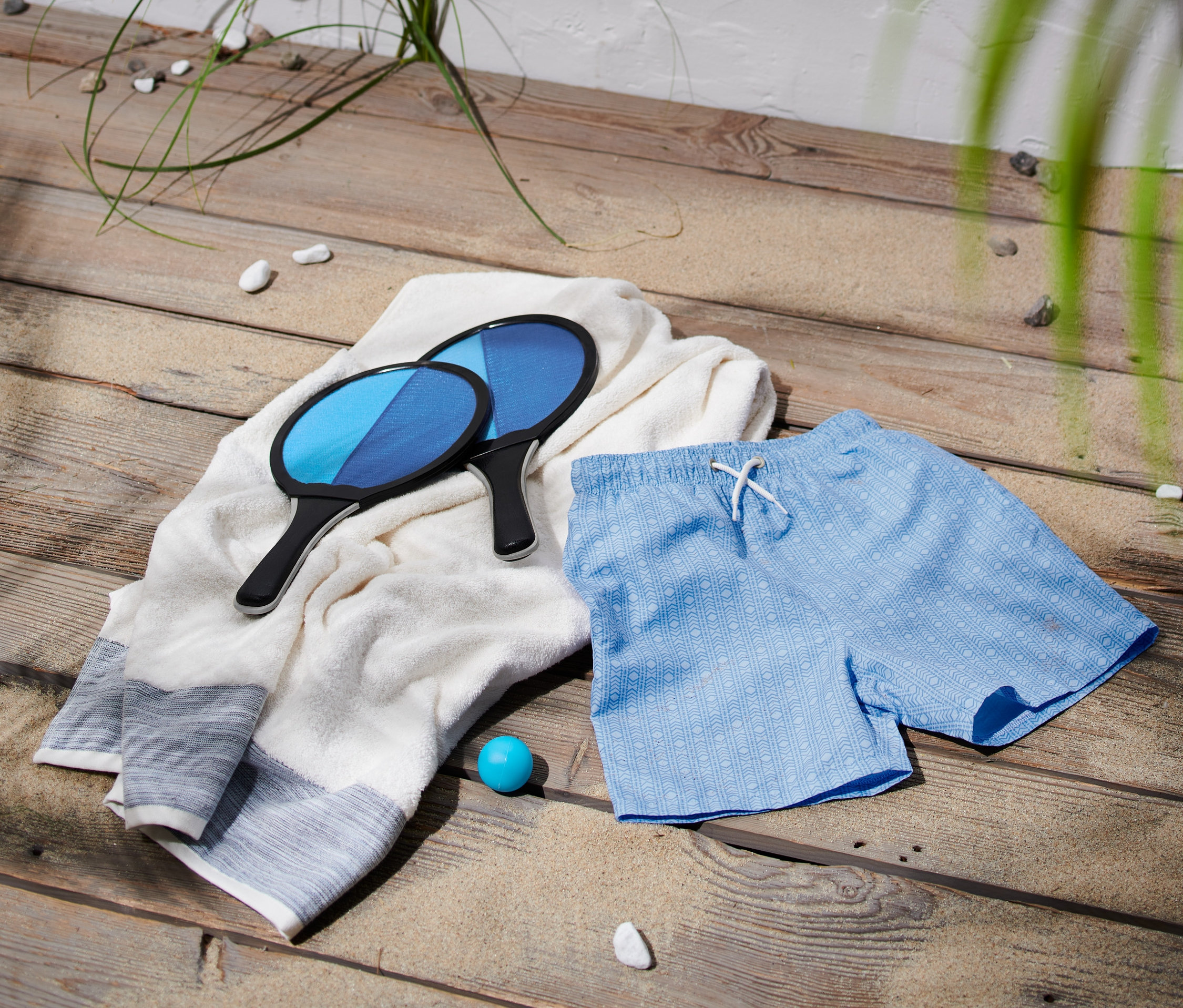 Zwei Badschläger liegen auf einem weißen Handtuch neben blauen Boardshorts aus recyceltem Polyester.