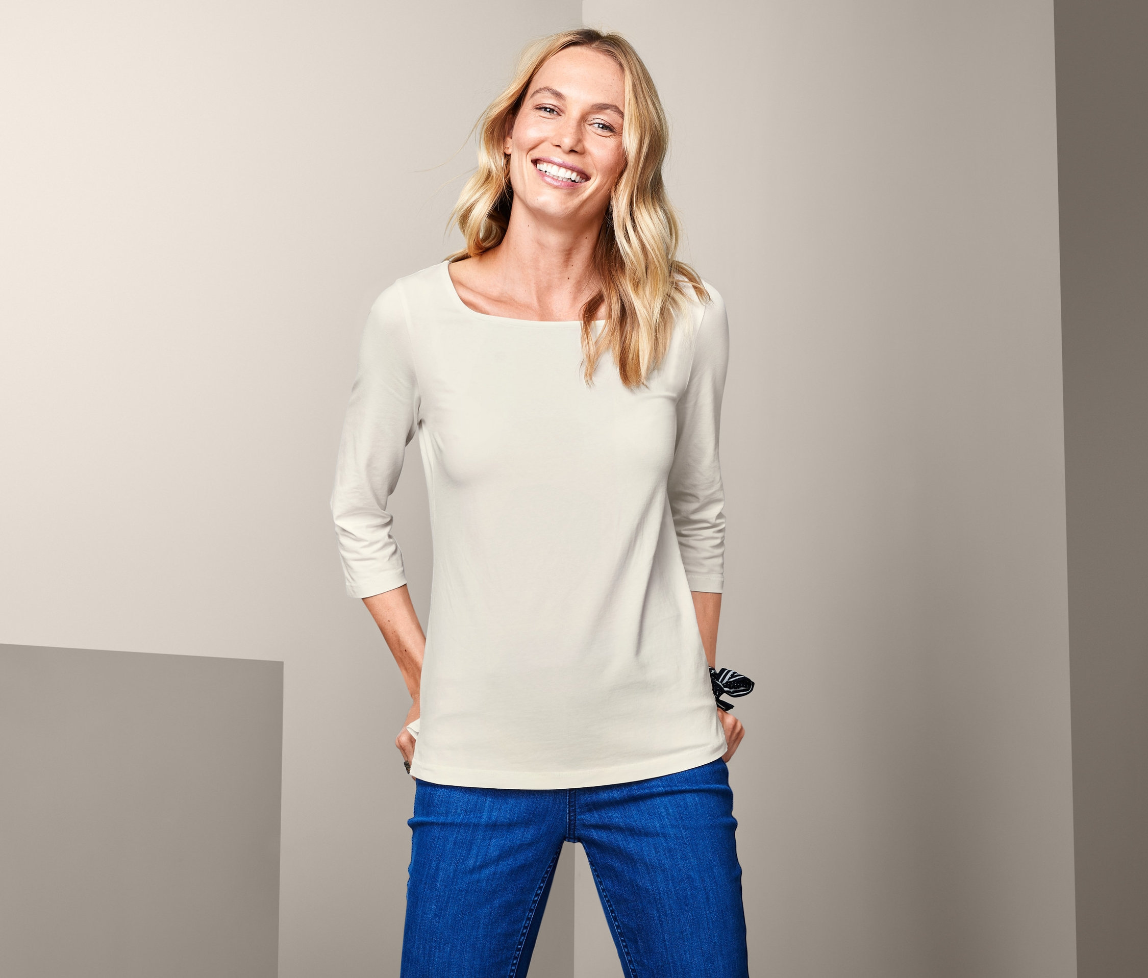 Frau mit blonden Haaren posiert in einem cremefarbenen Shirt mit 3/4-Arm und blauen Jeans.