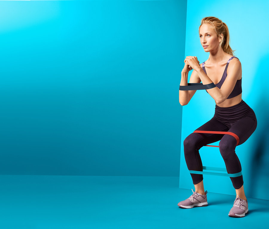Frau macht Kniebeugen mit 3 Trainingsbändern, eines um die Oberschenkel und eines um die Arme, vor blauem Hintergrund.