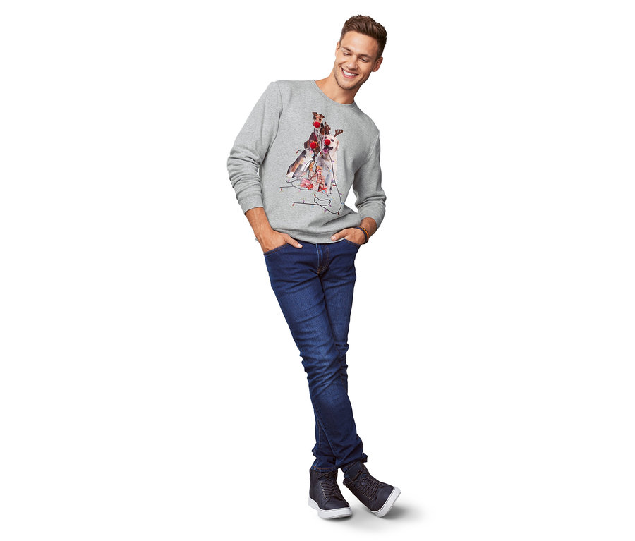 Mann in grauem Sweatshirt mit Hundeaufdruck und Jeans steht mit den Händen in den Hosentaschen.