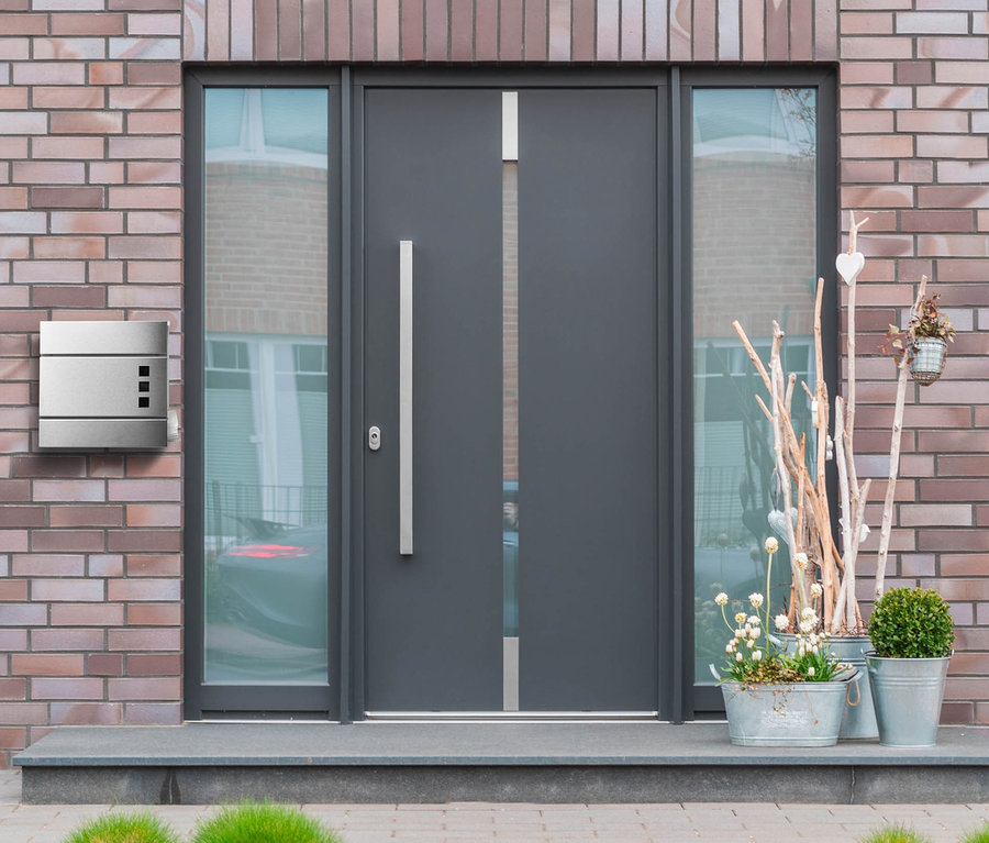 Graue Haustür mit Fenstern und ein Briefkasten mit Zeitungsfach und Sichtfenster an einer Backsteinmauer.