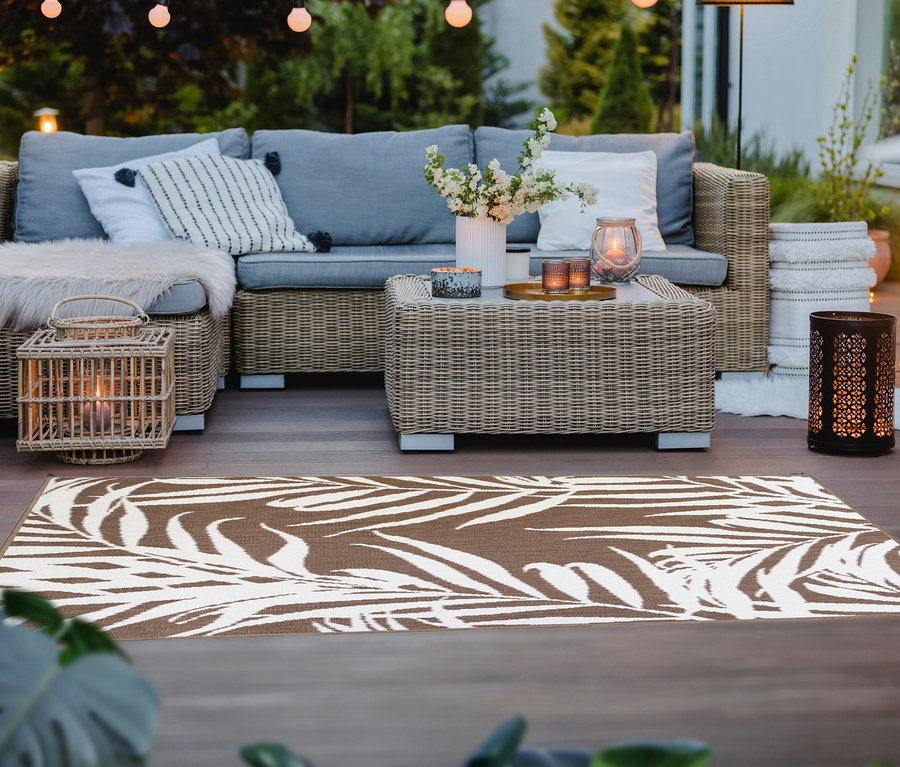 Ein brauner Outdoor-Wendeteppich »Palm Tree« (ca. 160 x 220 cm) liegt auf einer Terrasse mit Rattanmöbeln.