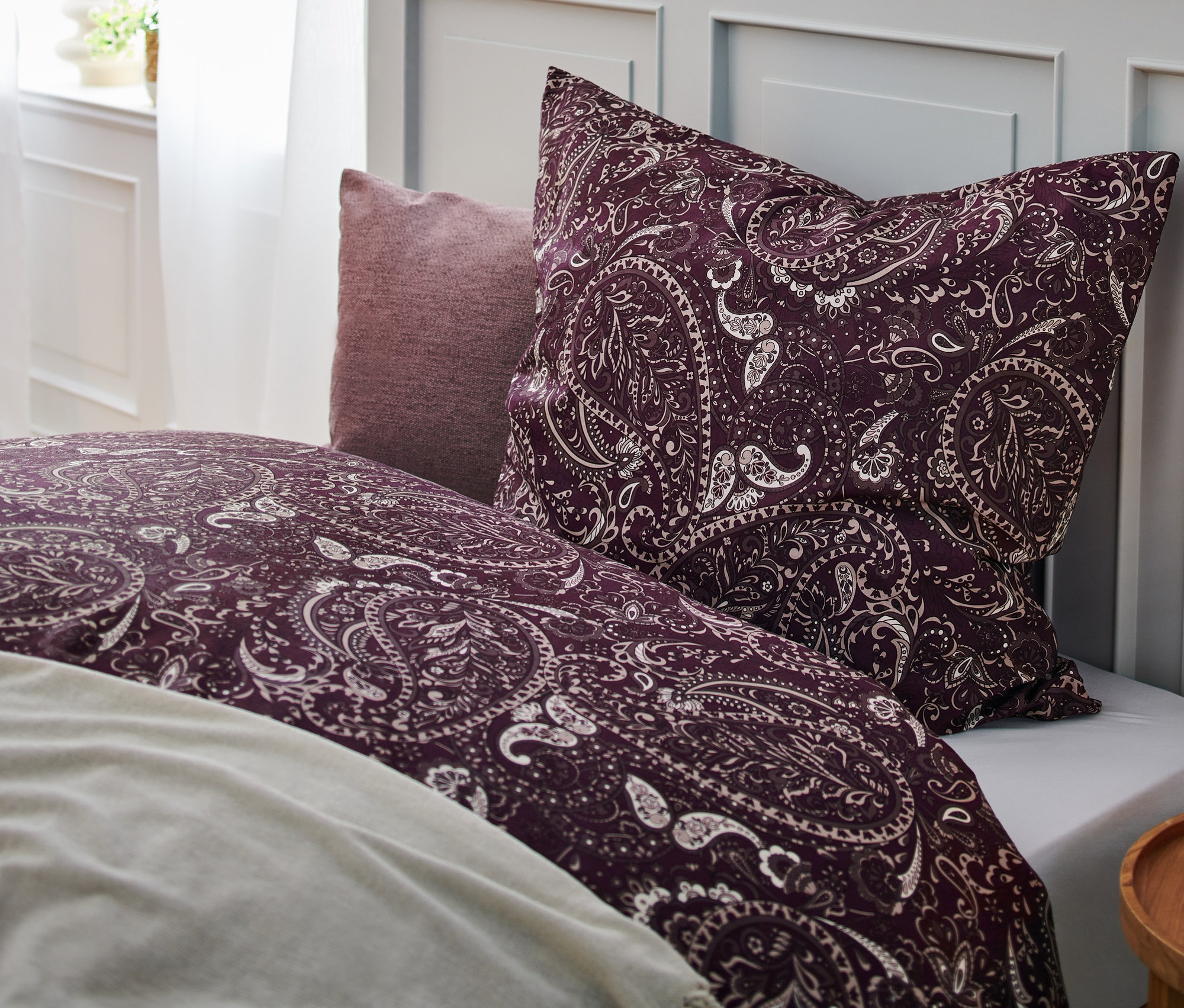 Nahaufnahme eines Bettes mit lila Bettwäsche und Kissen mit Blumenmuster und einem Nachttisch mit einer brennenden Kerze. Sichtbar sind eine Renforcé-Bettwäsche in Übergröße, ein Dekokissen-Bezug in Flieder und eine Renforcé-Bettwäsche in Normalgröße.