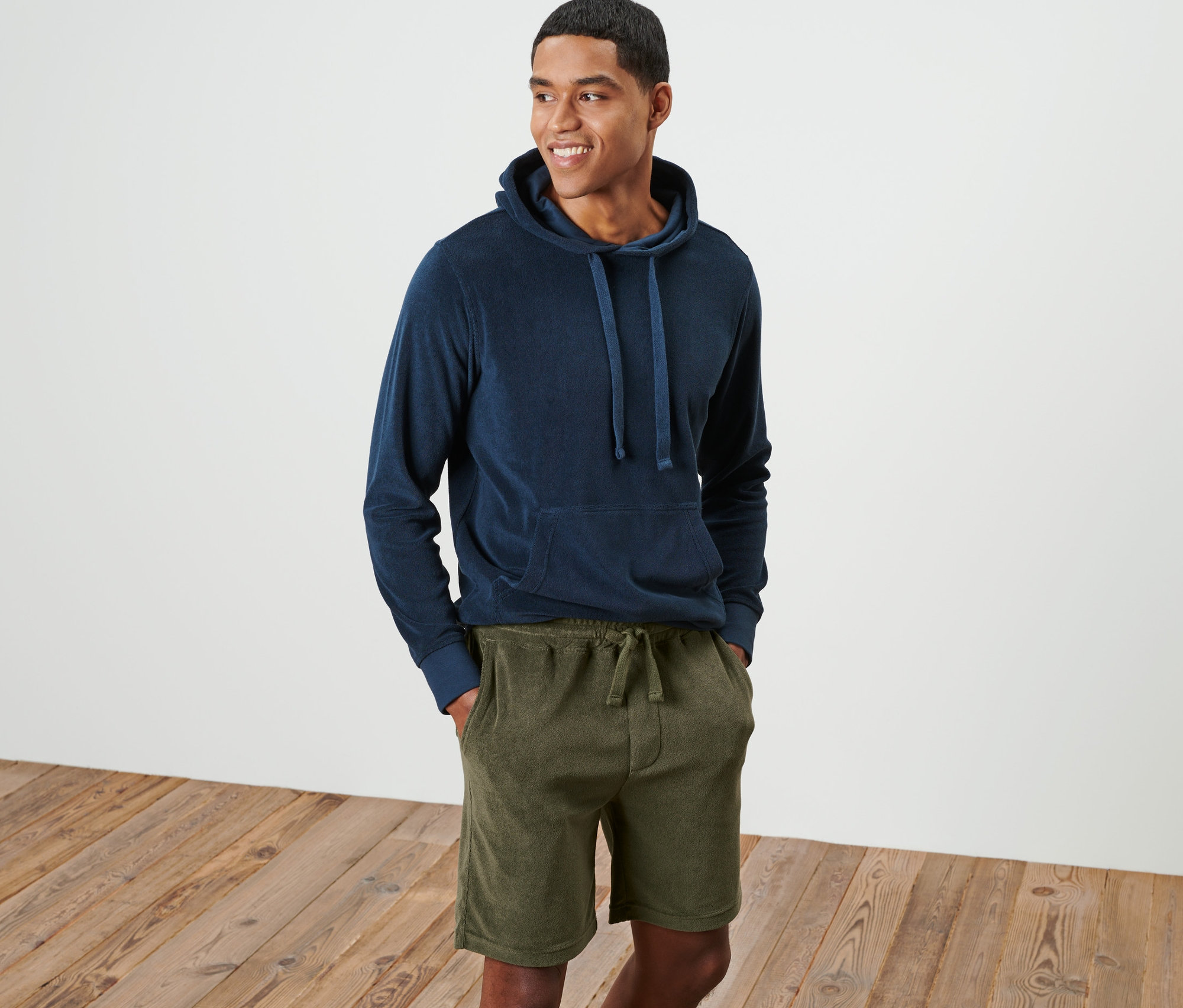 Ein lächelnder Mann steht und trägt einen blauen Kapuzenpullover und grüne Frottee-Shorts.