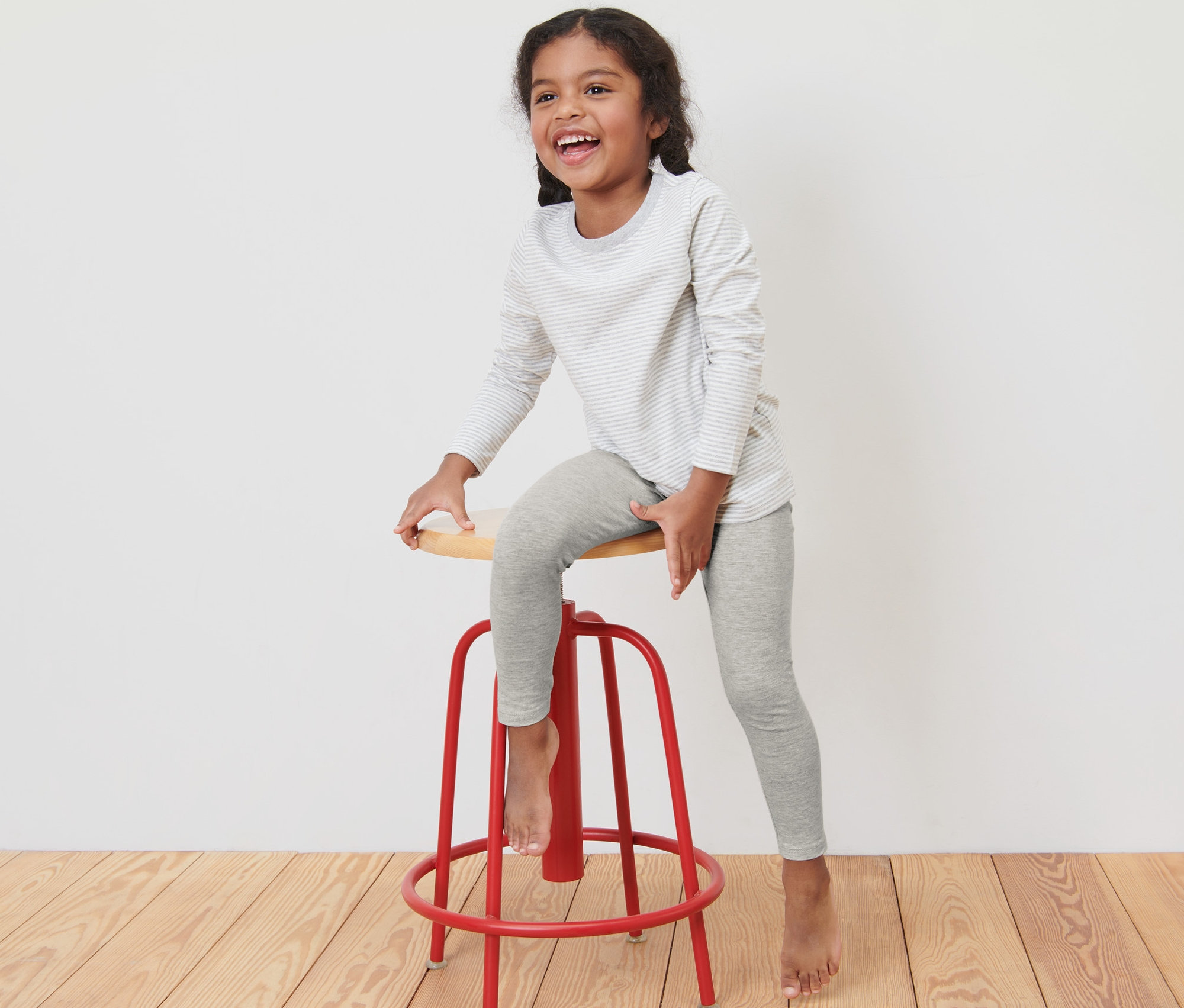 Ein Mädchen sitzt auf einem roten Barhocker und trägt graue Leggings und ein gestreiftes Langarmshirt.