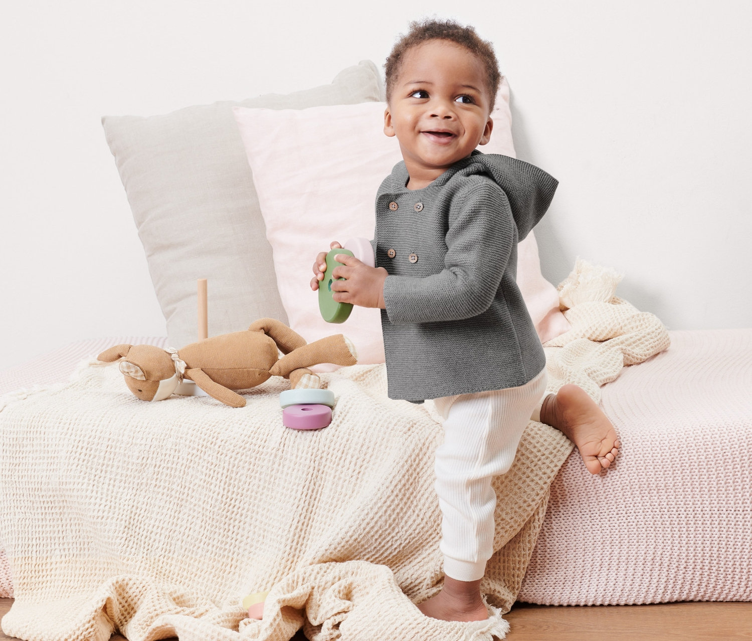 Ein Baby steht auf einem Bett und hält ein grünes Spielzeug. Es trägt eine grau melierte Baby-Strickjacke.