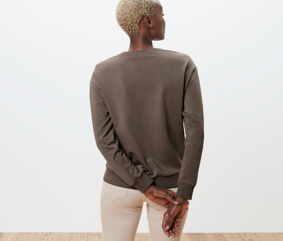 Rückansicht einer Frau mit kurzen blonden Haaren, die ein braunes Sweatshirt mit Stickerei und eine beige Hose trägt.