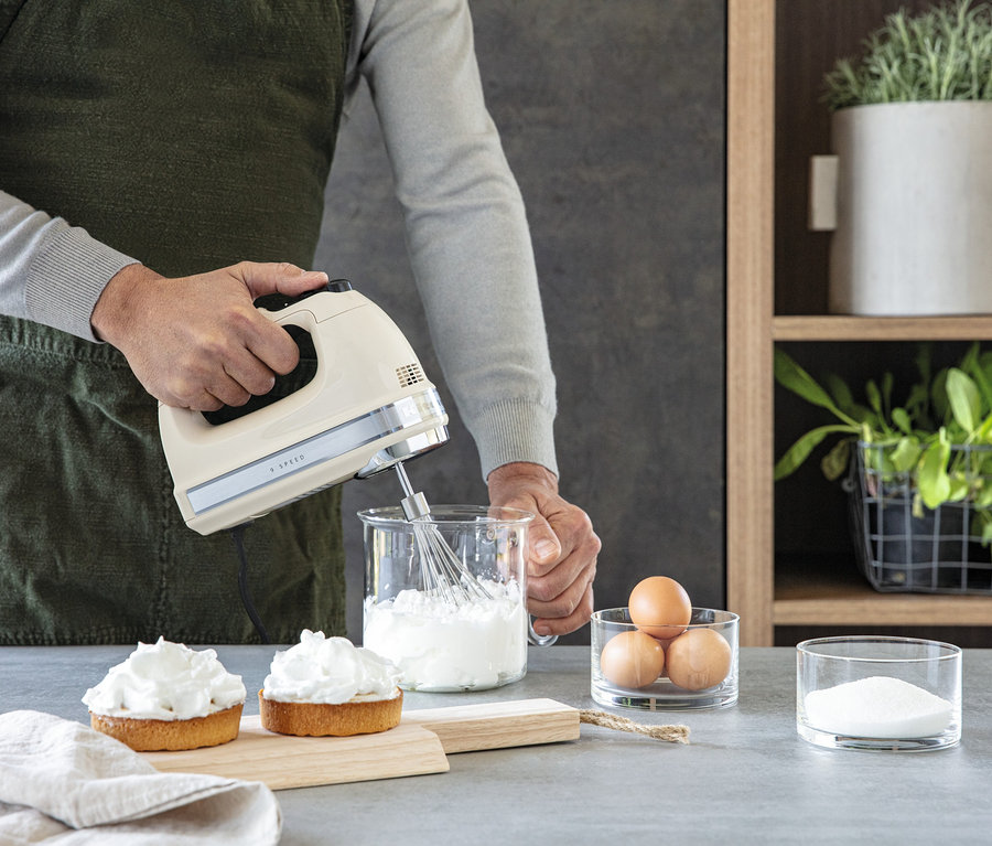 Mann mixt mit einem KitchenAid-Handmixer »5KHM9212EAC« Zutaten in einer Glasschüssel. Auf dem Tisch liegen zwei Törtchen, Eier und Zucker.