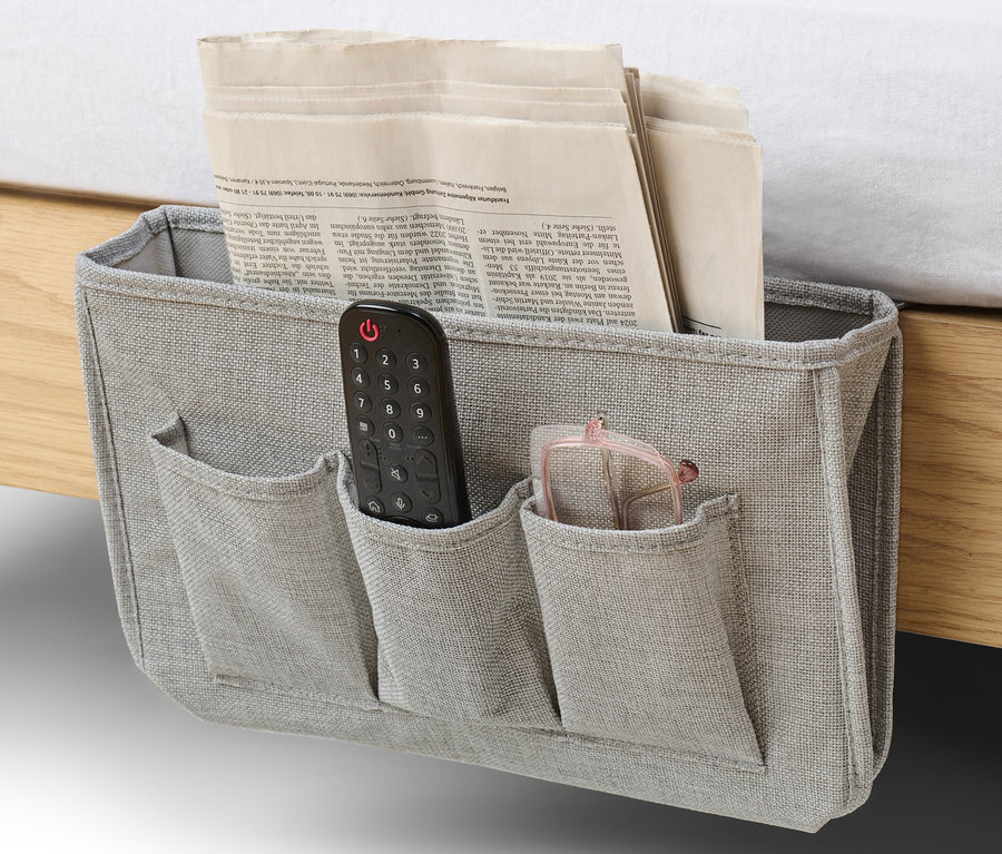Ein grauer Bett-und-Couchorganizer hängt an der Seite eines Bettes und enthält eine Zeitung, eine Fernbedienung und eine Brille.