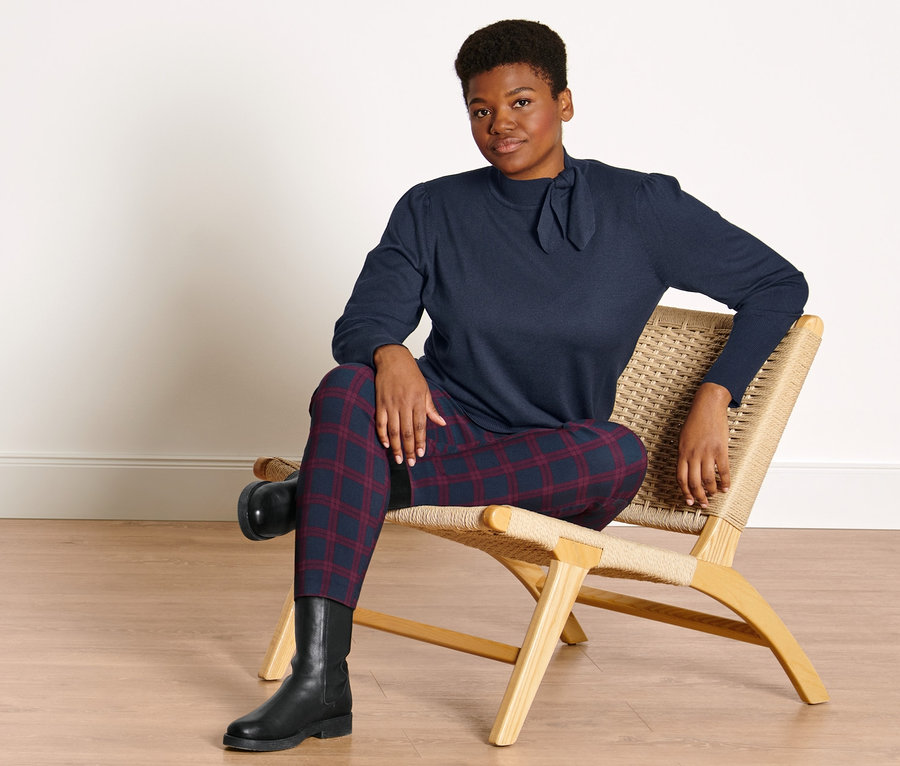 Frau sitzt auf einem Stuhl. Sie trägt einen Feinstrickpullover mit Schleifendetail, eine Stretchhose, rot-blau kariert, und Lederboots.