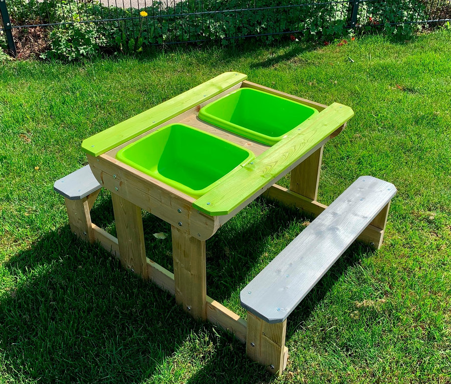 Auf einer Wiese steht ein Wendi Picknick- und Matschtisch mit Bänken und zwei grünen Plastikwannen.