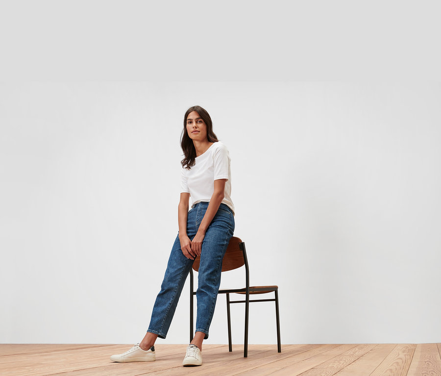 Frau sitzt auf einem Stuhl und trägt Mom Jeans – Fit »Nyla« und ein weißes T-Shirt.