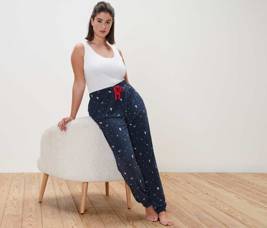 Eine Frau, die sich an einen flauschigen Hocker lehnt, trägt ein weisses Tanktop und eine Pyjamahose.