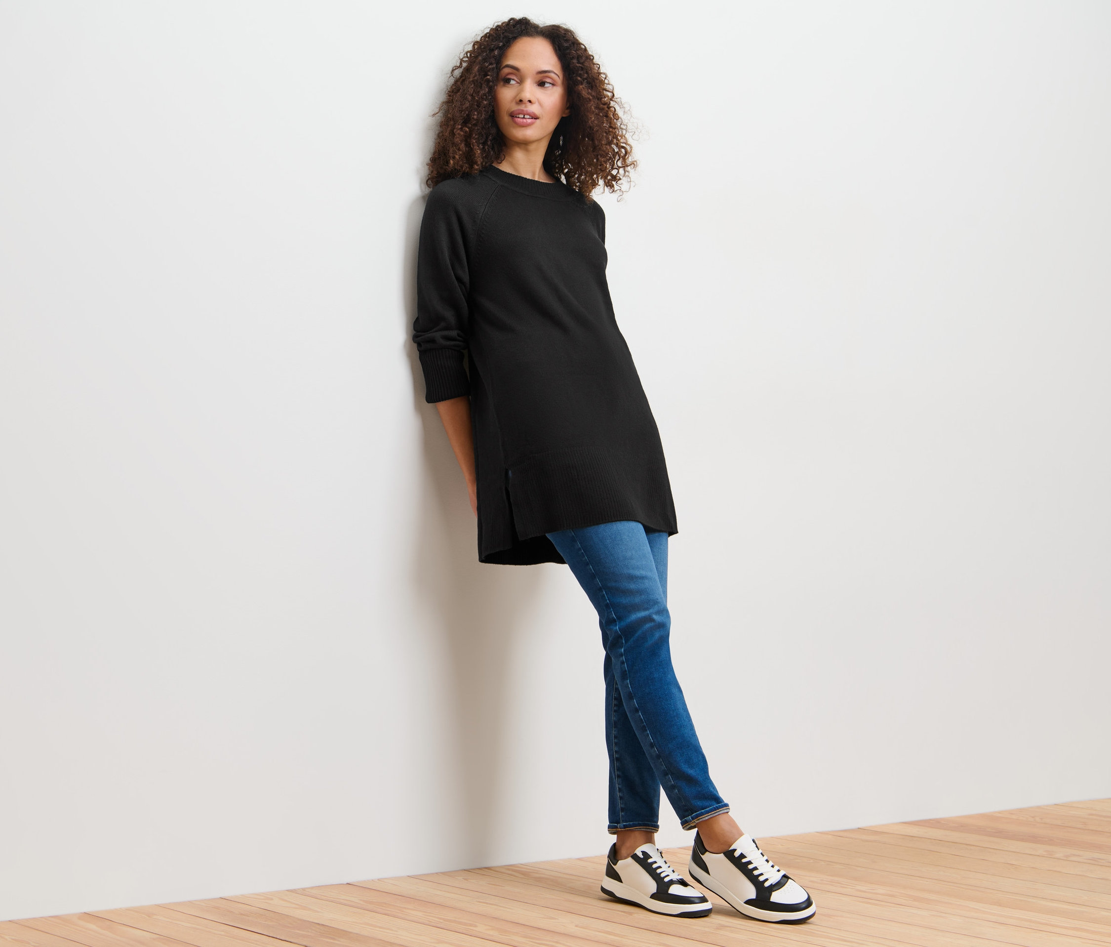 Frau lehnt an Wand, trägt langen schwarzen Strickpullover.