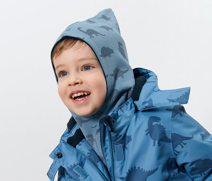 Ein Junge trägt eine Kinder-Wende-Schlupfmütze mit Dino-Motiv und eine Jacke mit Dino-Muster.
