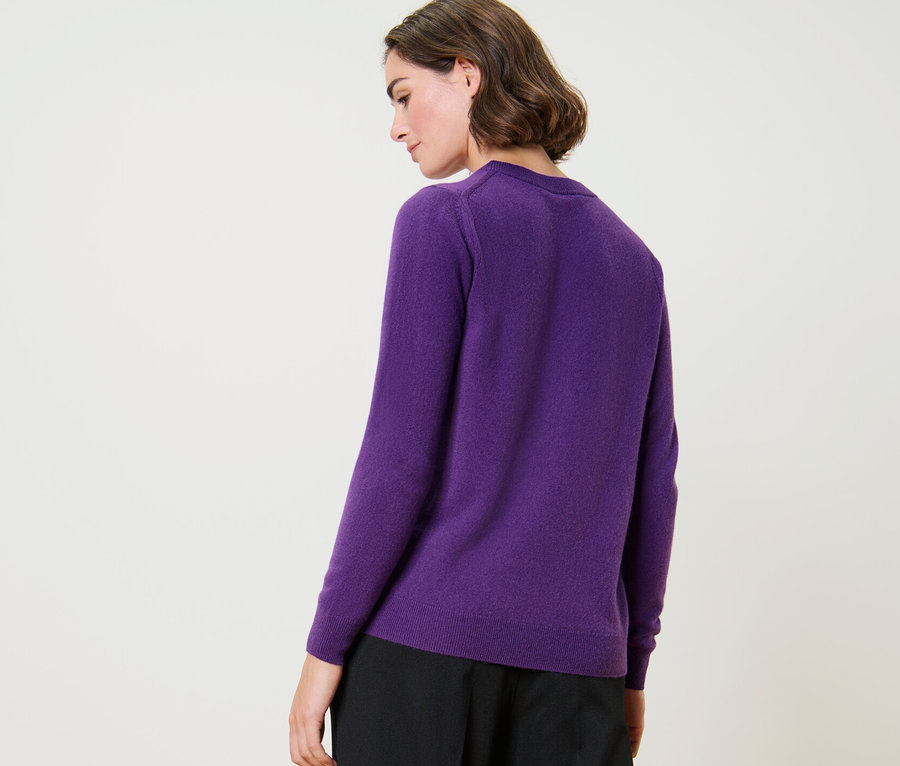 Frau posiert mit dem Rücken zur Kamera und trägt einen lila Feinstrickpullover mit Kaschmir und eine schwarze Hose.