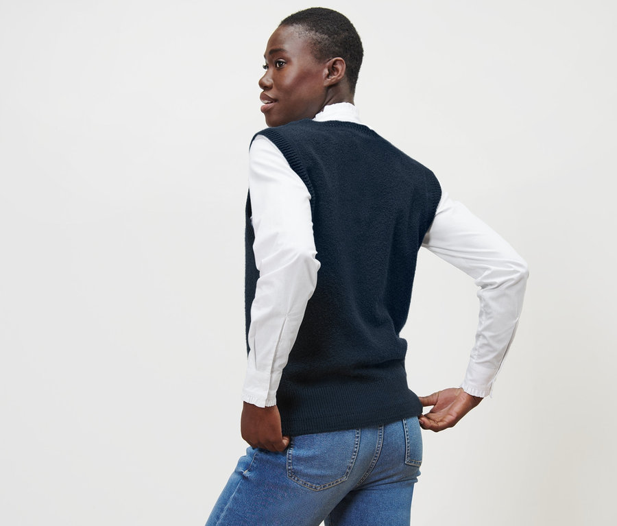 Frau mit Strick-Pullunder in Boucléoptik, weißem Hemd und blauen Jeans steht mit dem Rücken zur Kamera.