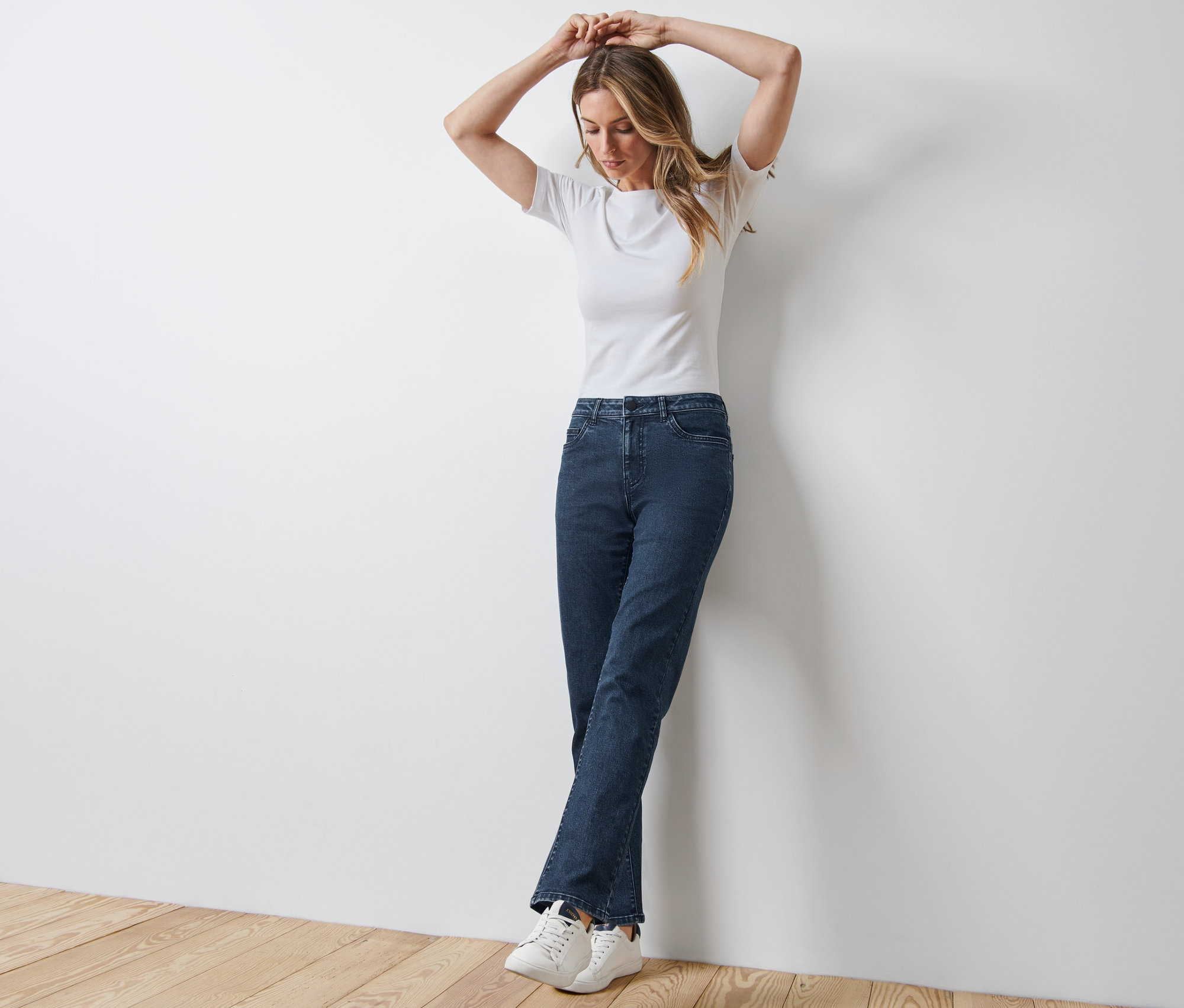 Eine Frau steht an einer weißen Wand mit ihren Händen über dem Kopf. Sie trägt Straight Jeans – Fit »Kira«, ein weißes T-Shirt und Turnschuhe.