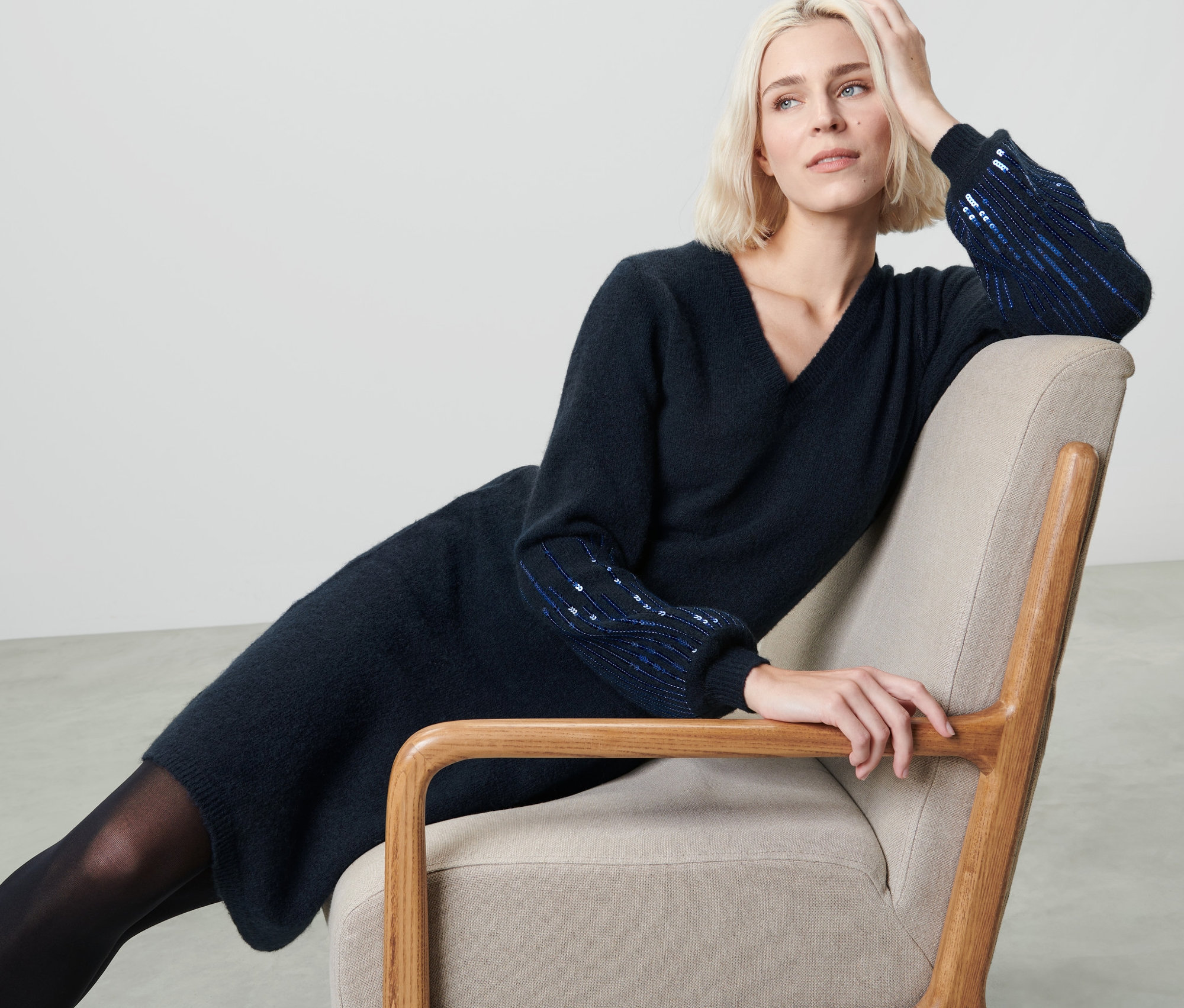 Blonde Frau sitzt im Sessel und trägt ein dunkelblaues Strickkleid mit Pailletten.