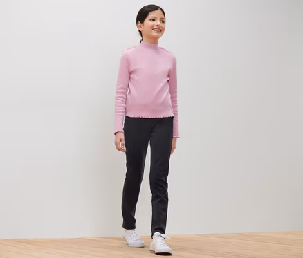 Lächelndes junges Mädchen posiert in einem hellrosa Oberteil und einer schwarzen Kinder-Treggings – Fit »Jule«.