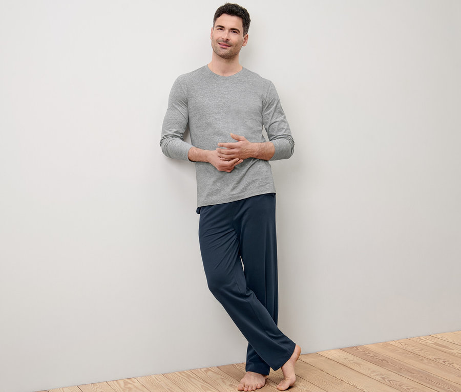 Mann lehnt an Wand, trägt grauen Pyjama und blaue Hose.