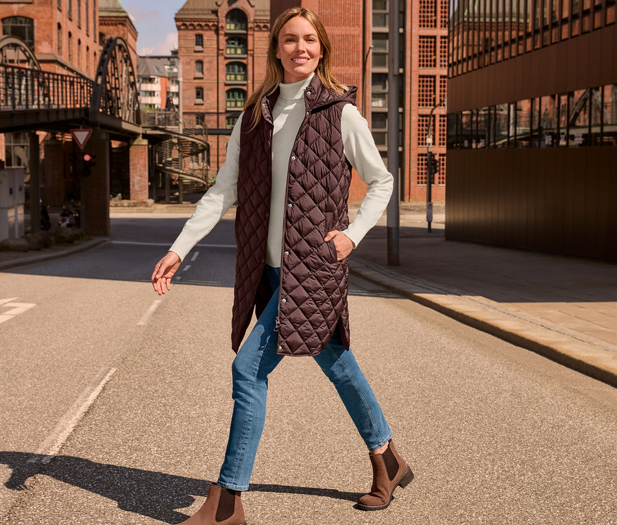 Eine Frau geht die Straße entlang und trägt eine lange Steppweste mit Kapuze, einen cremeweißen Feinstrick-Rollkragenpullover, eine Slim Jeans – Fit »Emma«, mid blue, und Veloursleder-Boots.