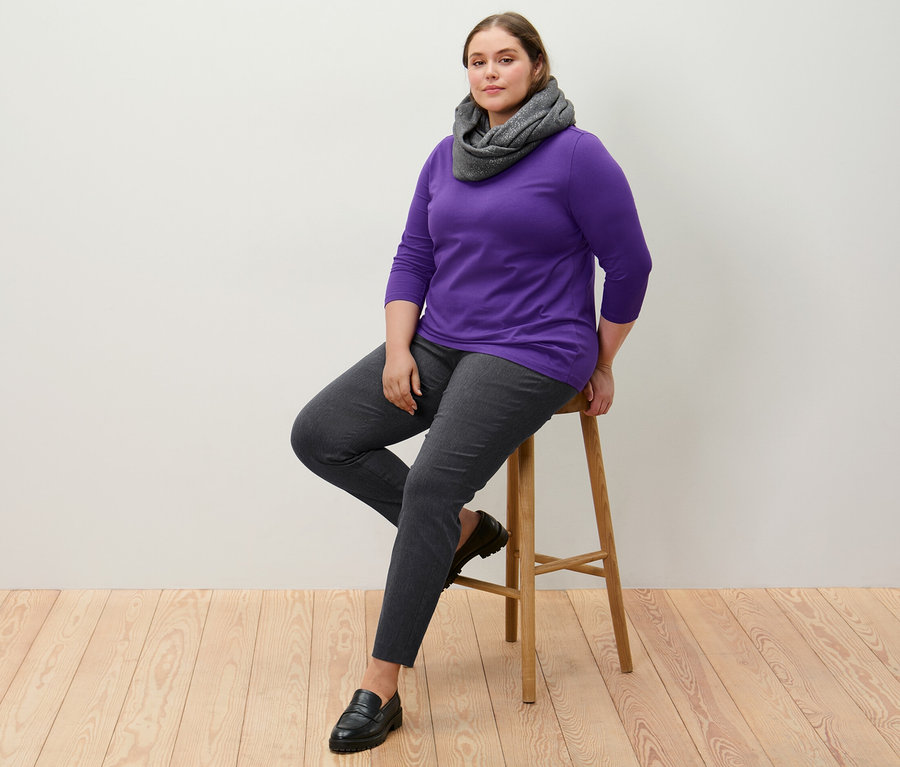 Frau sitzt auf einem Hocker, trägt eine Stretchhose und ein lila Shirt mit 3/4-Arm.